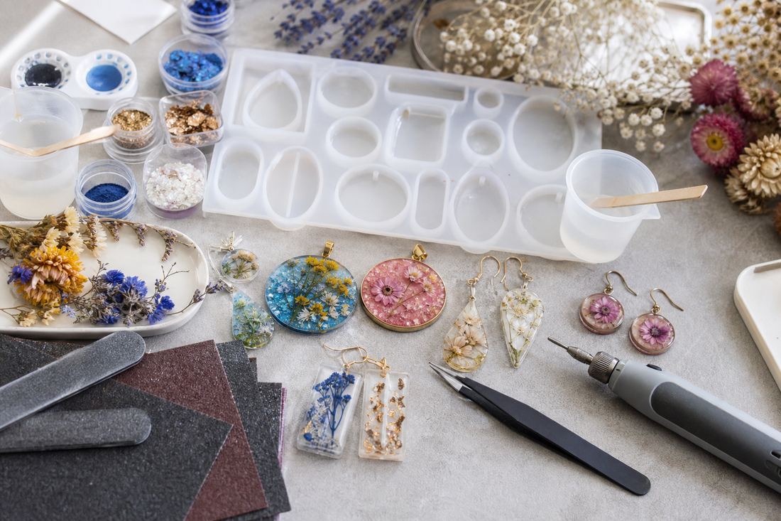 Finished resin pendants and earrings arranged with molds, pigments, dried flowers, and finishing supplies on a craft workspace