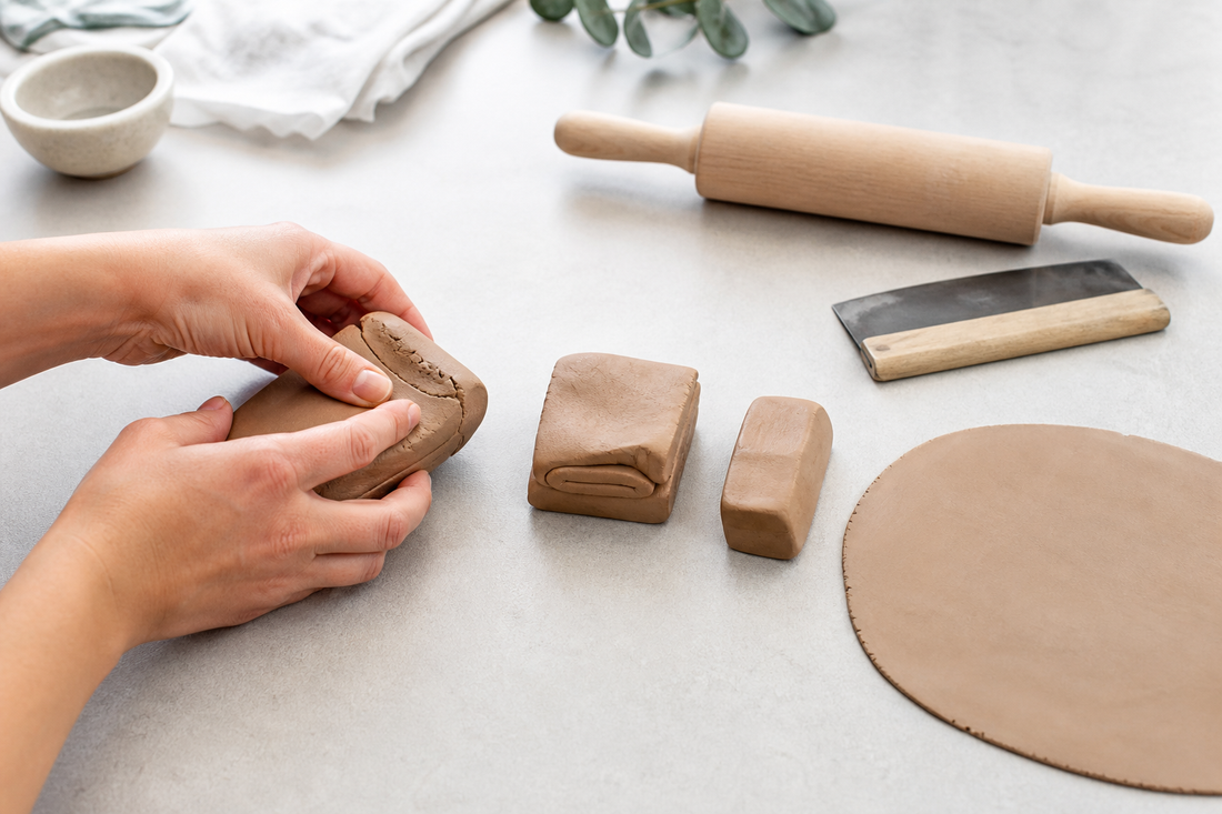 Hands conditioning polymer clay beside a rolling pin, with a smooth sheet and small clay pieces on a clean work surface