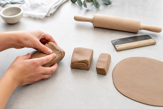 Hands conditioning polymer clay beside a rolling pin, with a smooth sheet and small clay pieces on a clean work surface